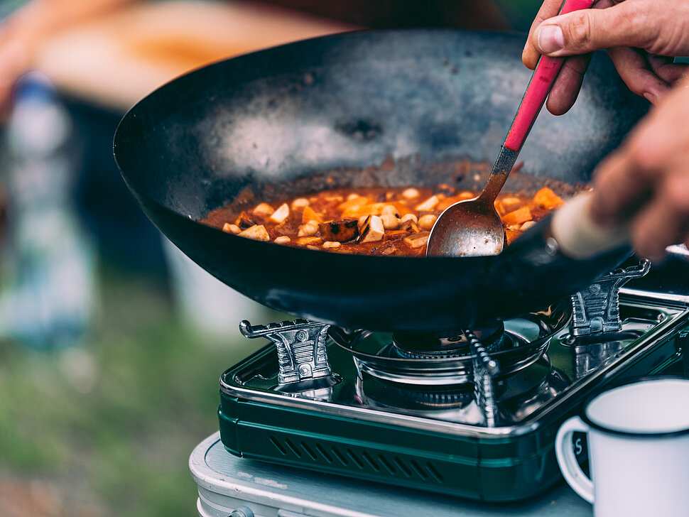 Hand rührt ein Gericht in einer Wok-Pfanne auf einem Camping-Herd